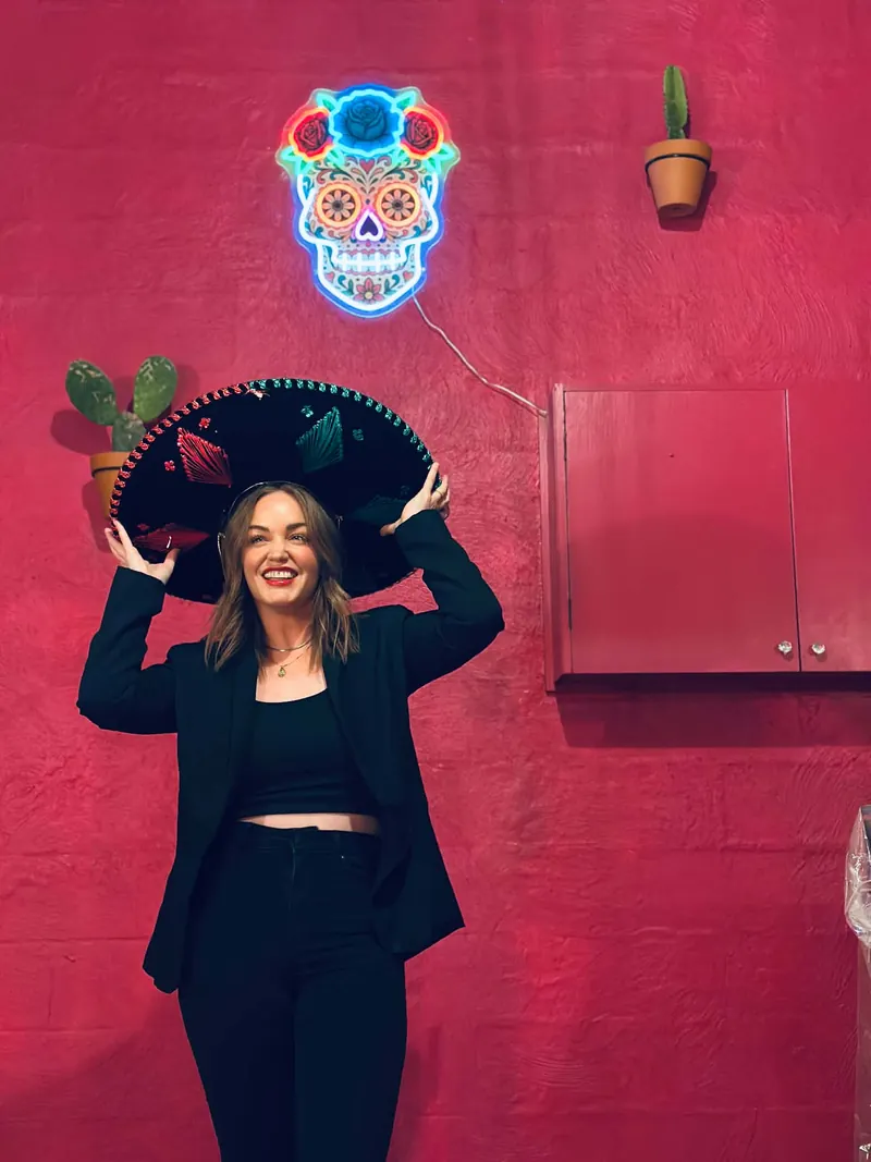 A smiling guest holding a sombrero beneath the neon skull